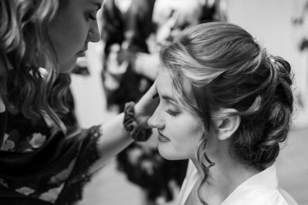 Black and white photo of makeup artist carefully applying cosmetics to bride with elegant updo hairstyle.