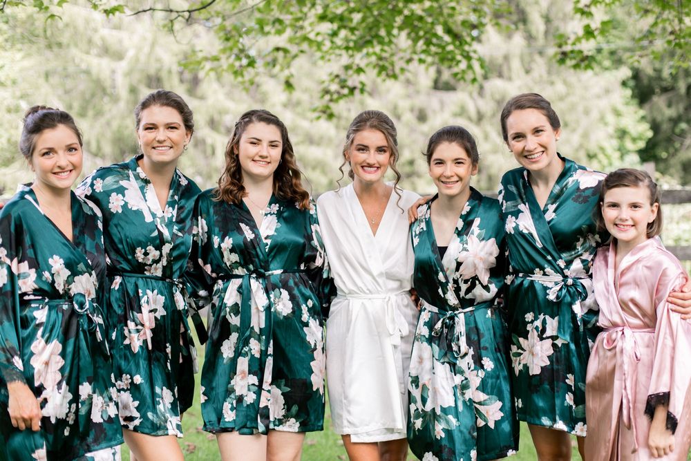 Bride in white robe poses with five bridesmaids in green floral robes and one in pink robe outdoors.