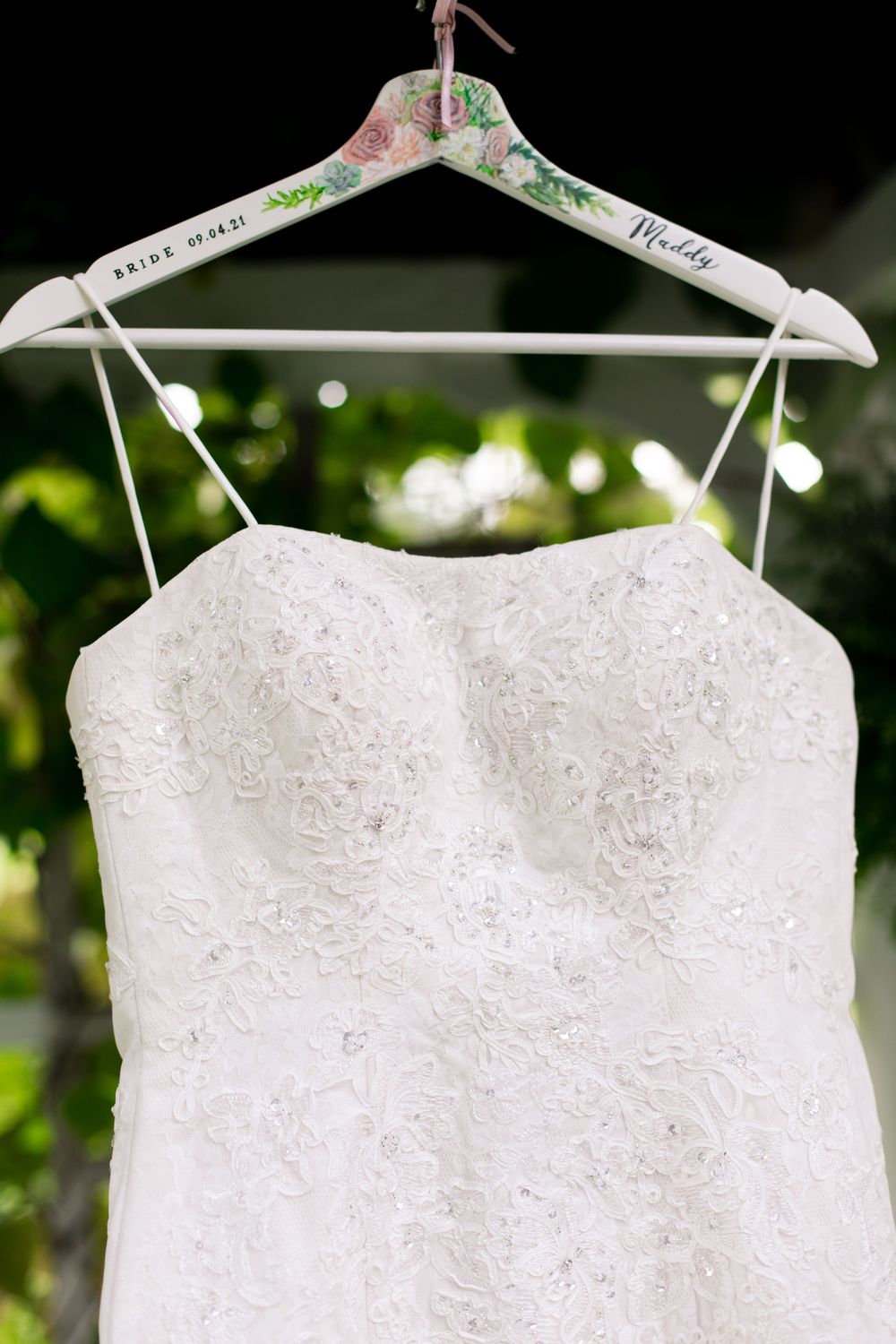 White lace strapless wedding dress hanging on a floral personalized bridal hanger outdoors.