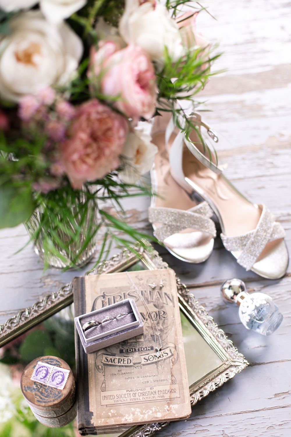 Vintage wedding flat lay with bridal bouquet, heels, rings, perfume, and sacred songs book.
