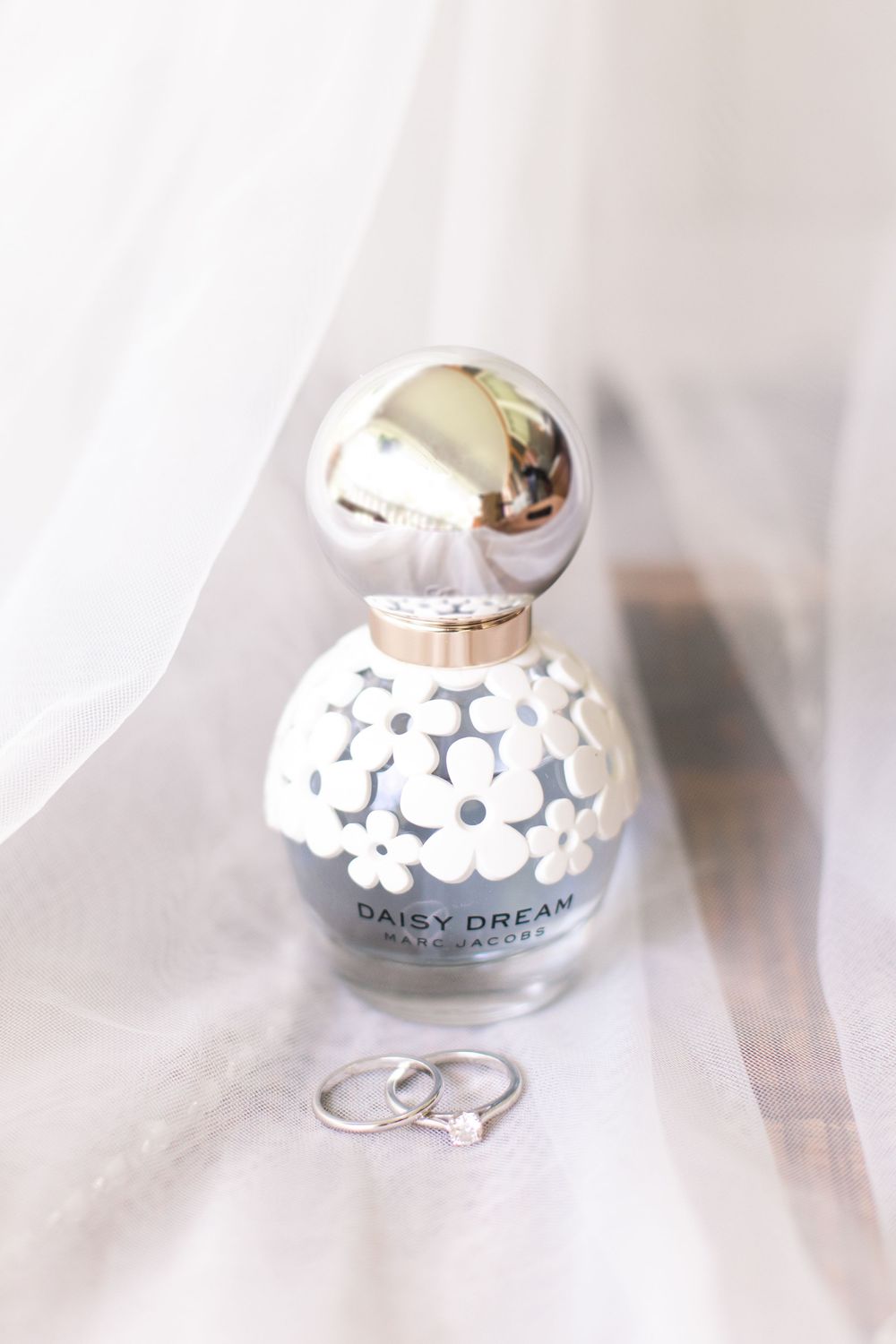 Marc Jacobs Daisy Dream perfume bottle with wedding rings on white bridal veil fabric.