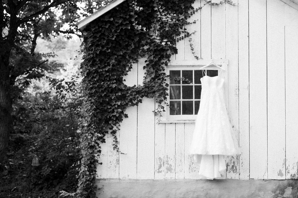 Black and white photo of elegant wedding dress hanging outside ivy-covered white barn.