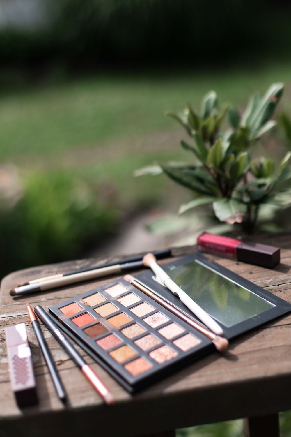 Eyeshadow palette with makeup brushes and cosmetics on a rustic wooden table outdoors.