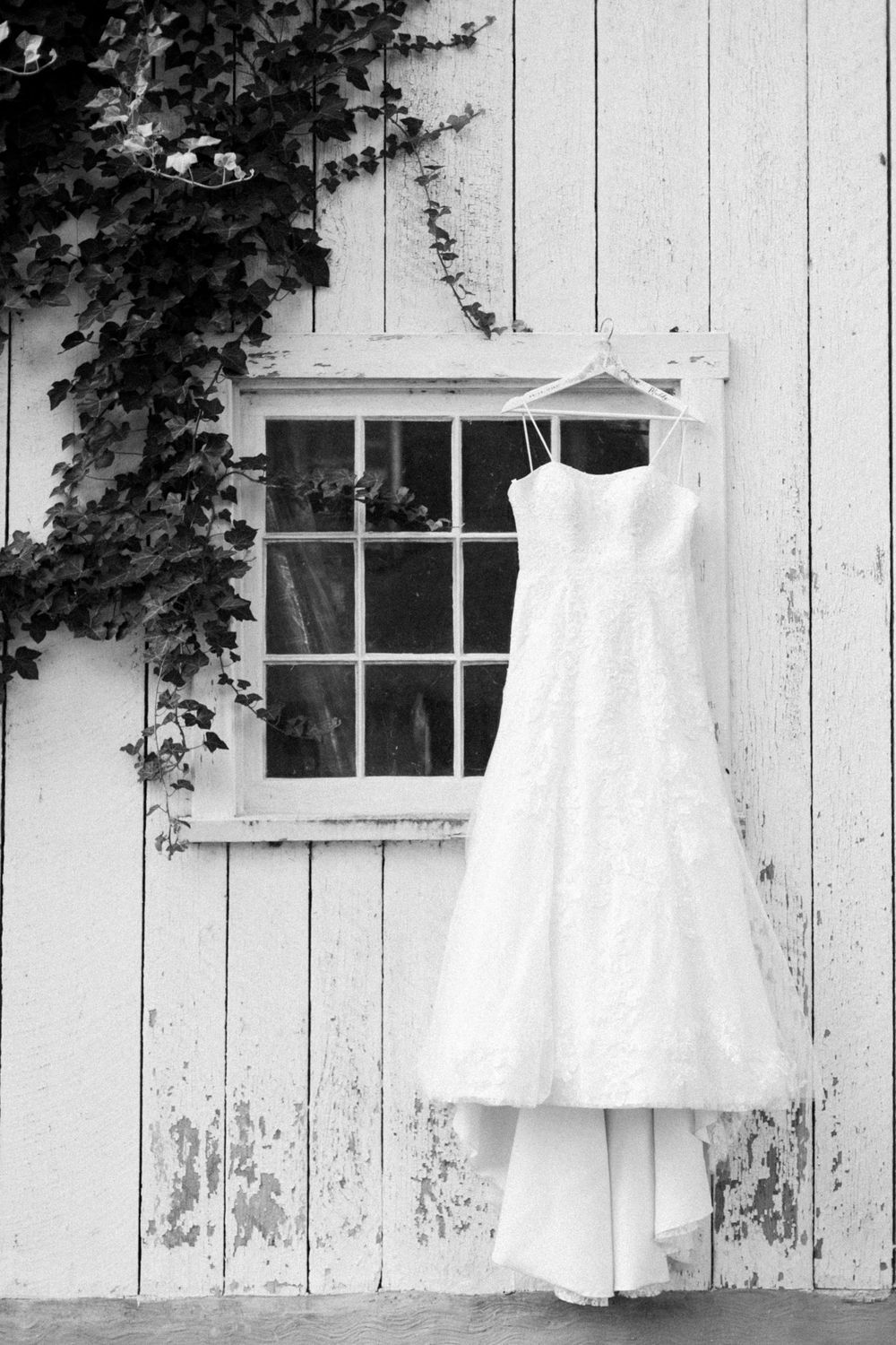 Black and white image of lace wedding gown hanging on rustic white barn window.