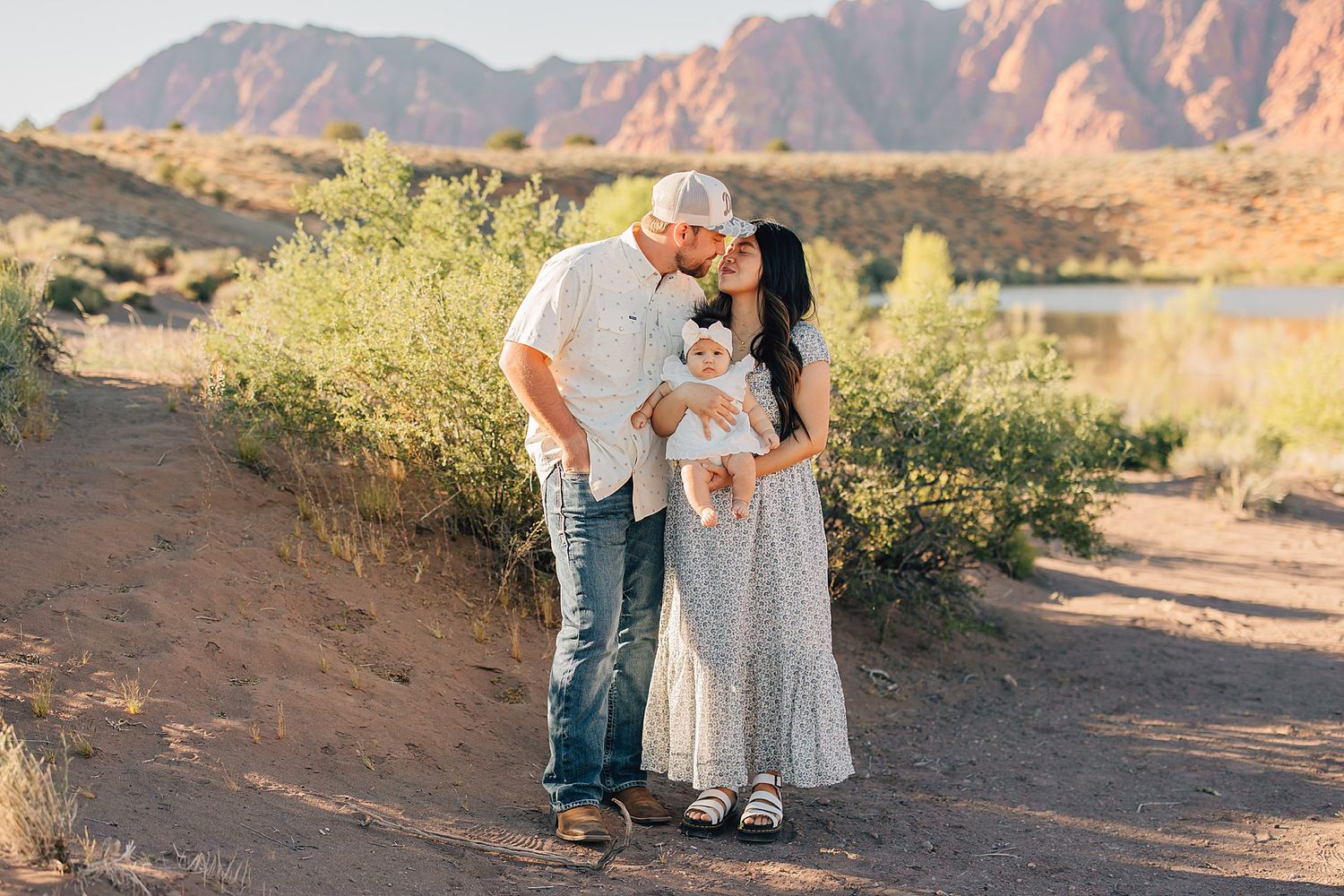 A couple kisses their baby outdoors on a desert trail with red rock mountains and a lake in the background.