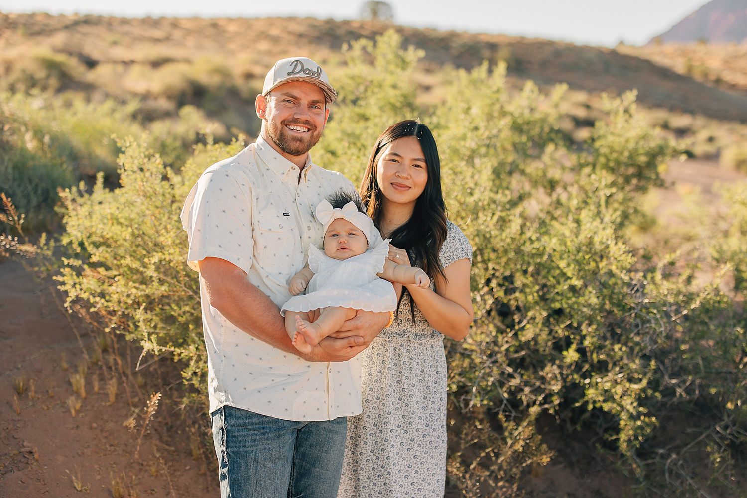 Couple holding newborn baby in desert landscape with warm sunset lighting and green shrubs.