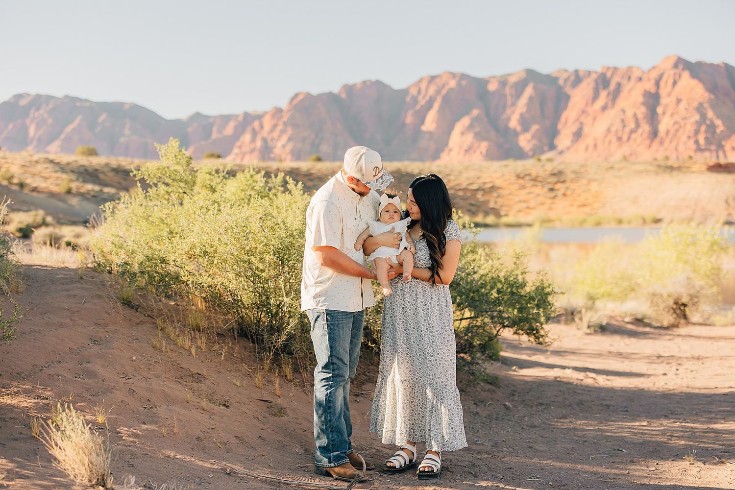 Family of three sharing a tender moment in a desert landscape with red rock mountains and a river in the background.