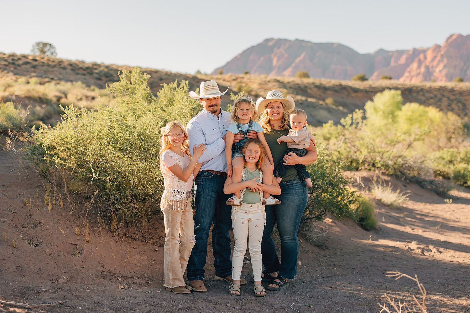 Family of six poses together in a scenic desert landscape with red rock mountains at golden hour.