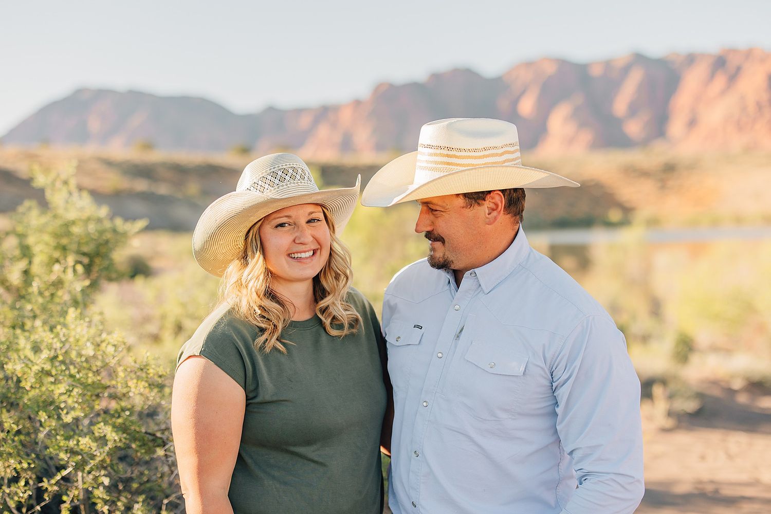 Couple in cowboy hats smiling together outdoors with red rock mountains and desert landscape in background.