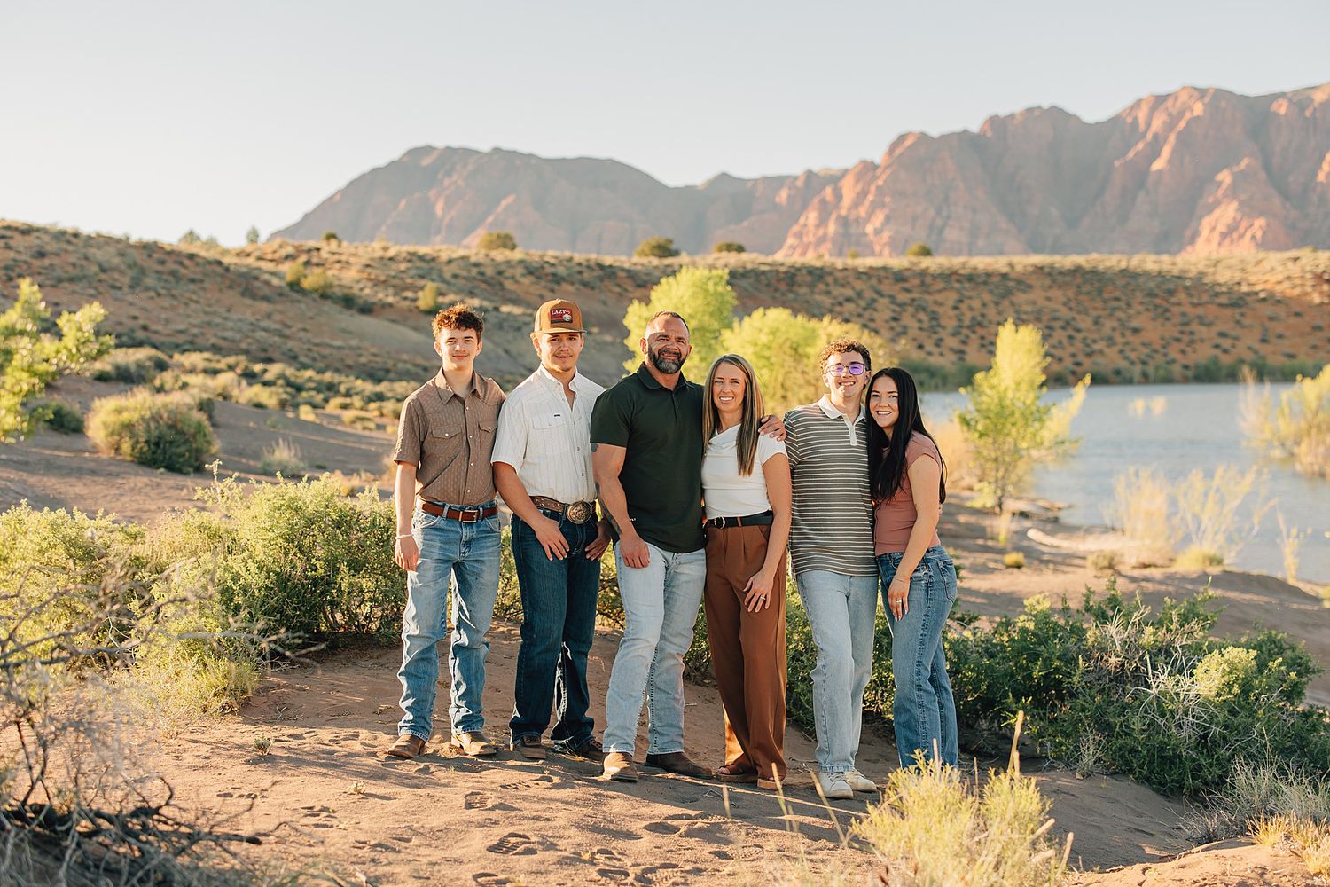 Six people smile together outdoors near a lake with red rock mountains in the background during golden hour.