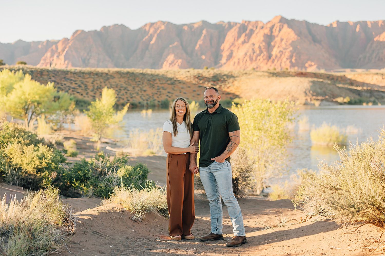 Couple smiling outdoors near a lake with red rock mountains in the desert background.