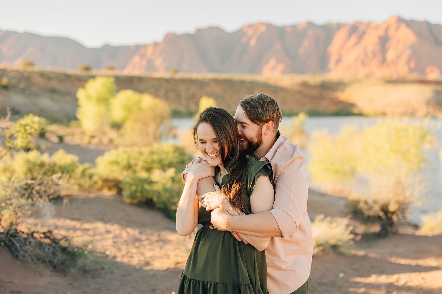 Couple embracing in desert landscape with mountains and river backdrop during golden hour engagement photo session.