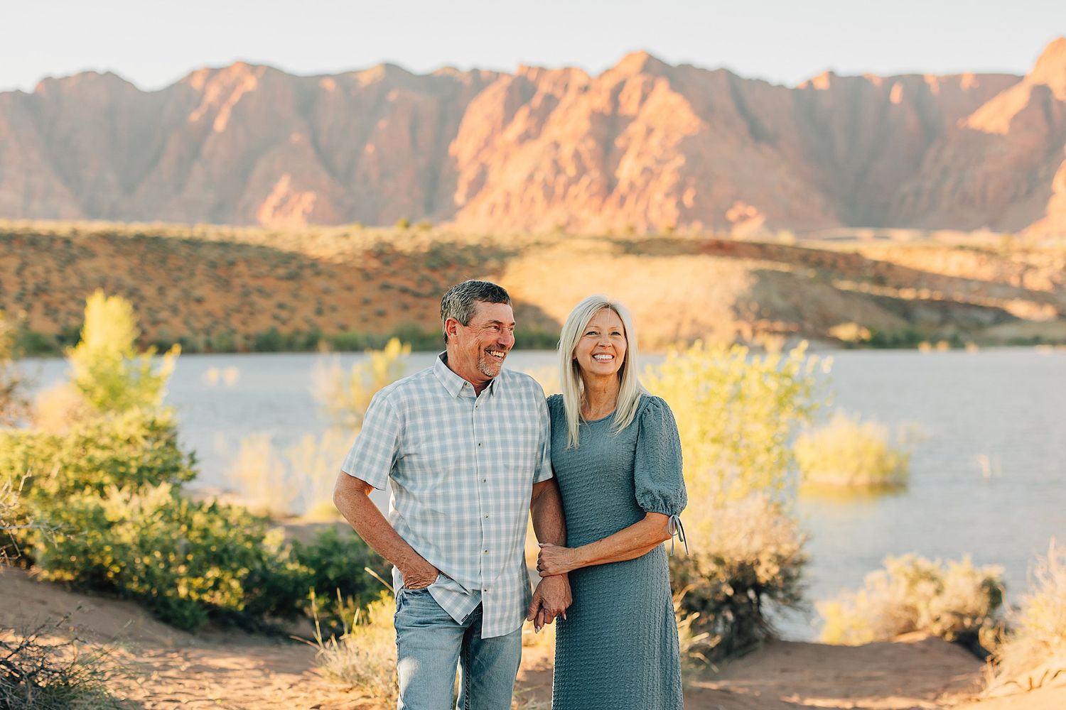 Smiling couple embracing outdoors with a scenic desert lake and red mountain backdrop in warm golden light.