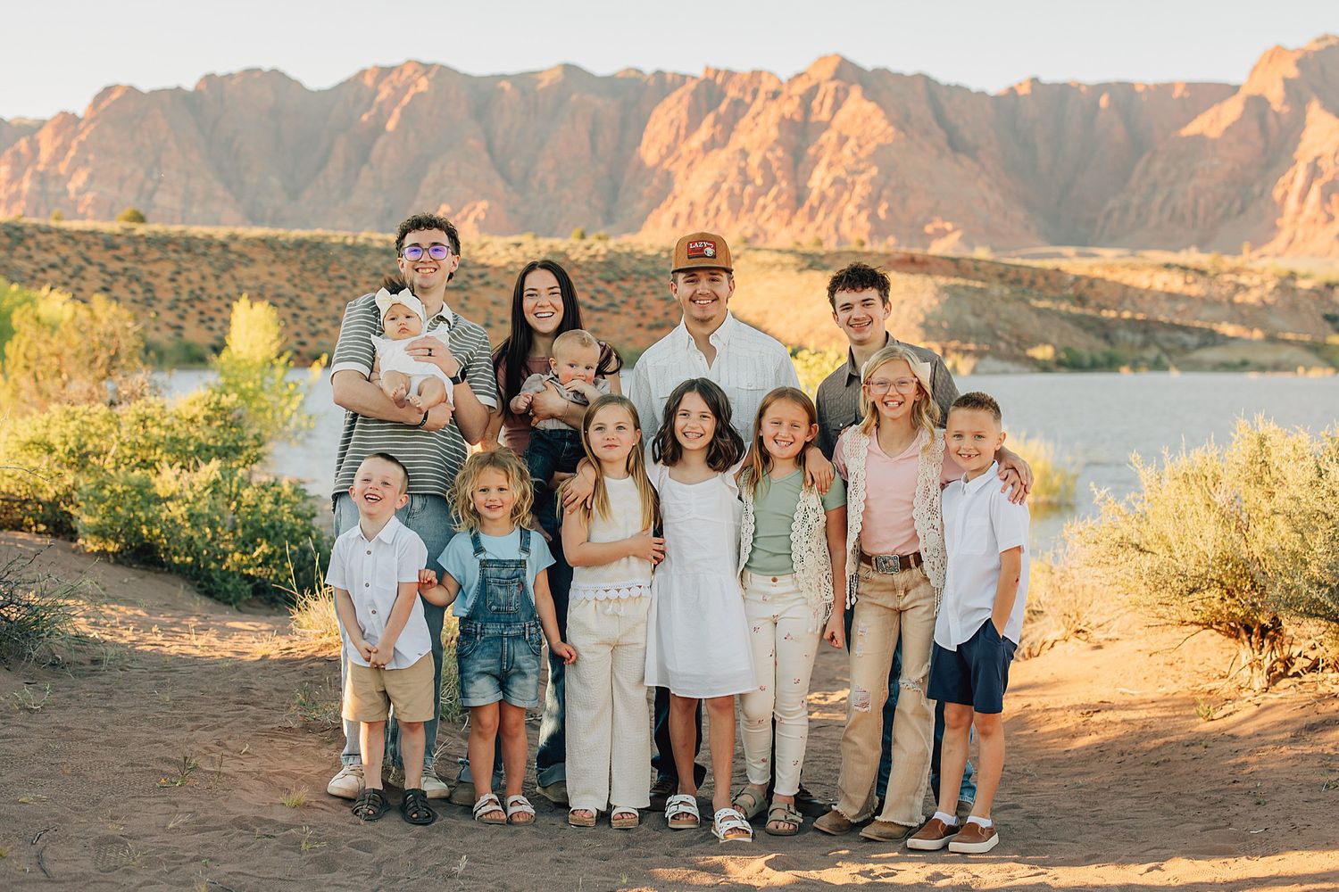 Large family of twelve posing outdoors near a lake with scenic desert mountains at golden hour.