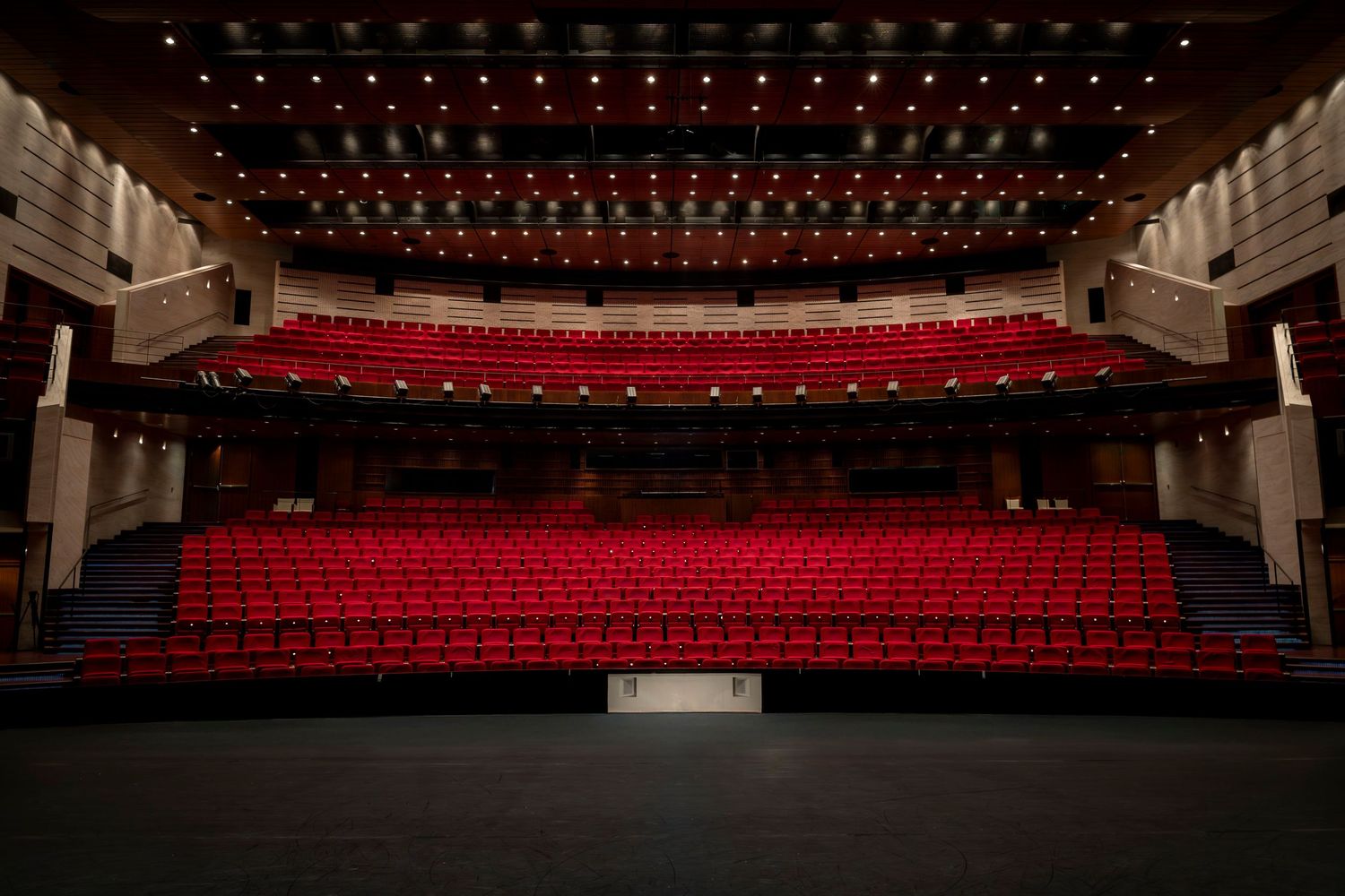 Empty red-seated theater auditorium viewed from the stage, with dramatic ceiling lighting above.