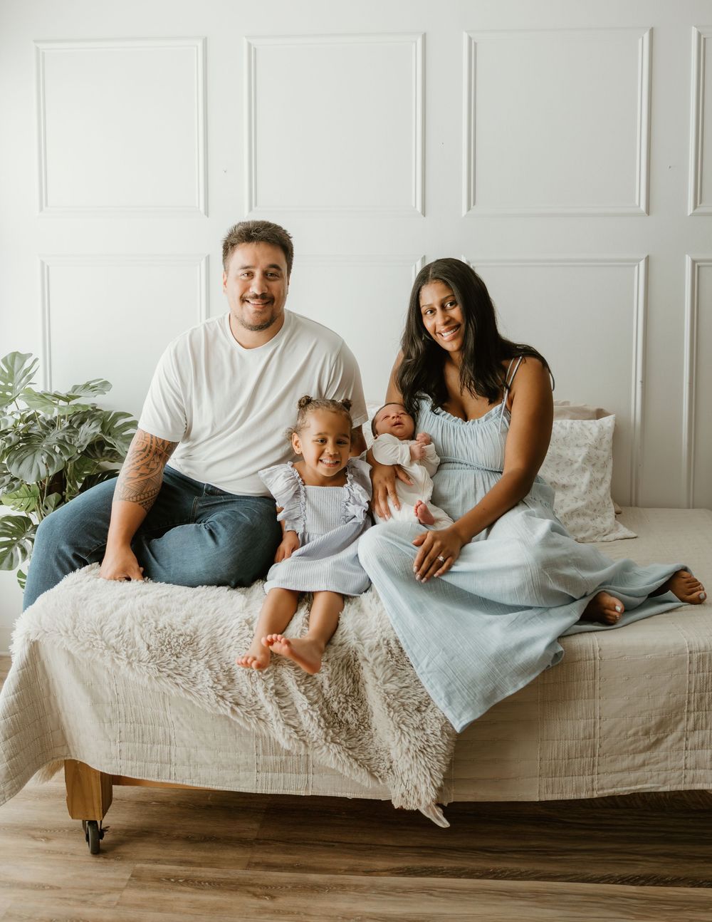Family of four with newborn baby posing on a bed, smiling warmly in a bright white studio setting.