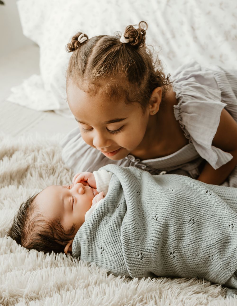 A young girl with curly pigtails smiles lovingly at a sleeping newborn swaddled in a light blue knit blanket.