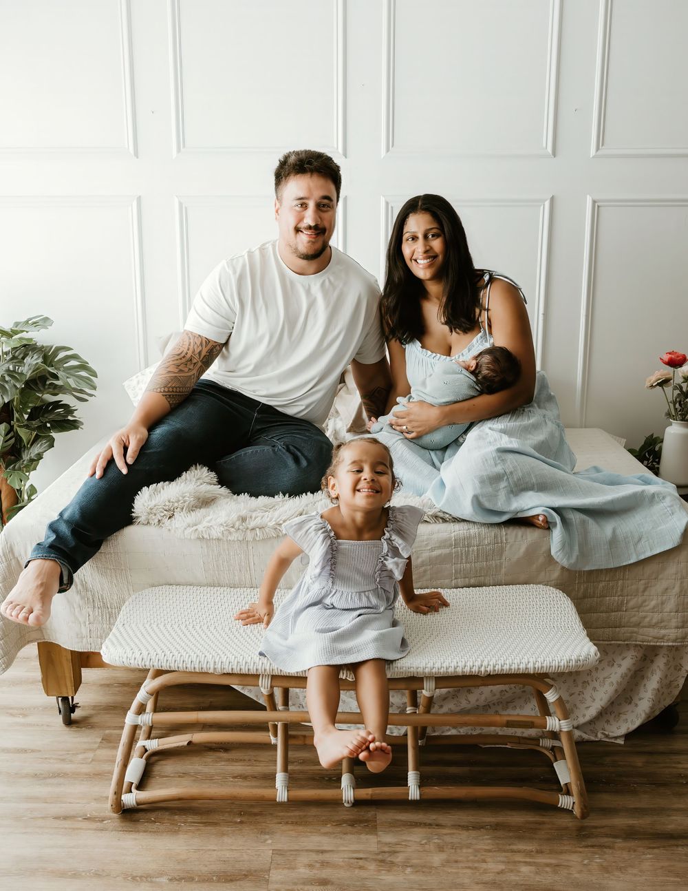 Happy family of four posing for a newborn photo session, with parents and toddler sitting on a white bed.