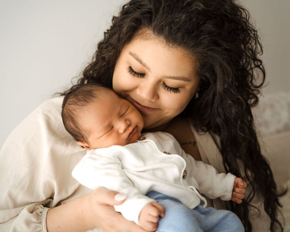 A mother with curly hair tenderly holds and cuddles her sleeping newborn baby dressed in white.