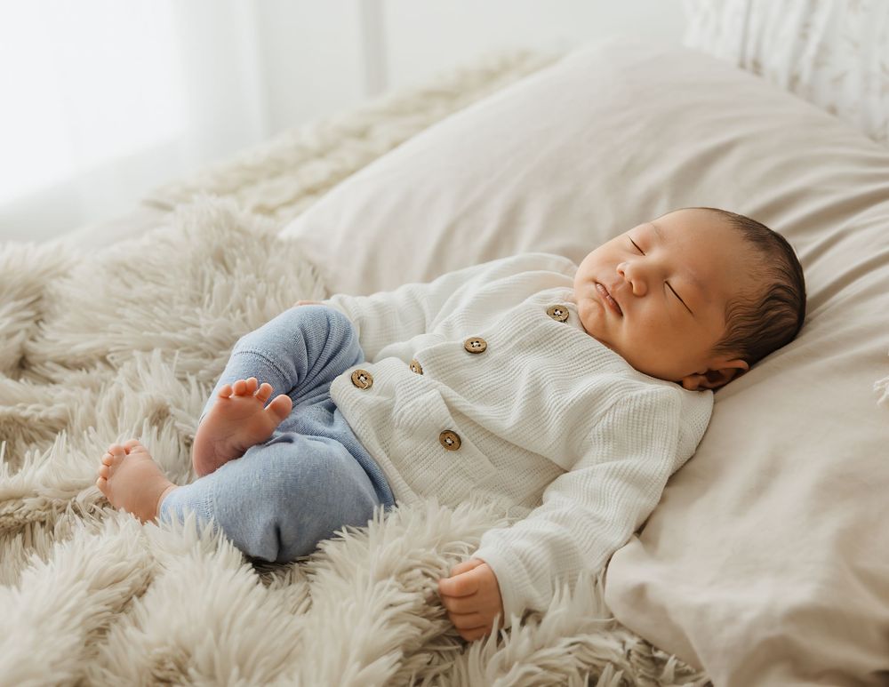 Peaceful newborn baby sleeping on a fluffy beige blanket wearing a white cardigan and blue pants.
