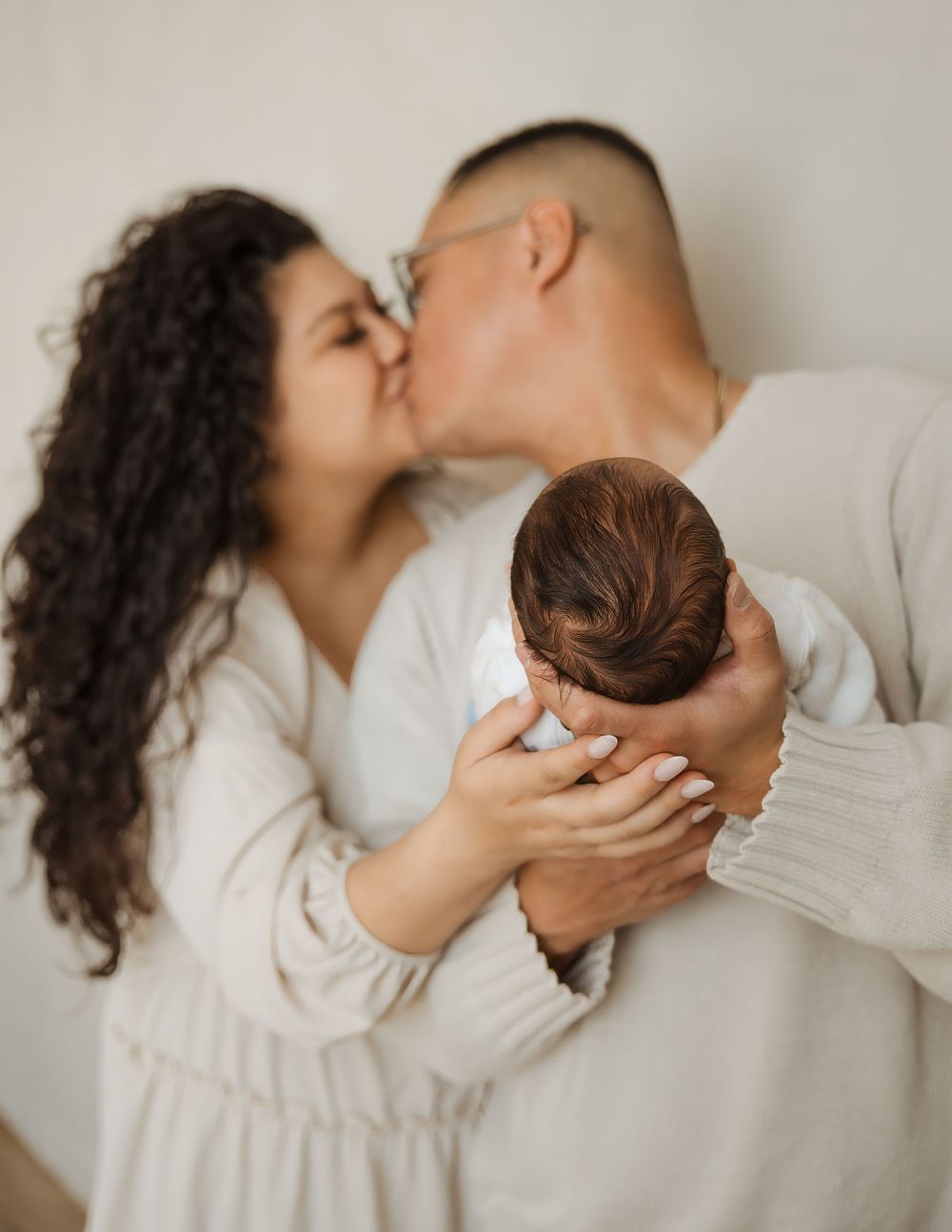 Parents share a kiss while cradling their newborn baby together in a bright, softly lit studio setting.