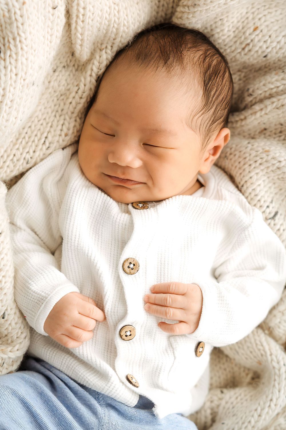 Peaceful newborn baby sleeping in white knit cardigan and blue pants on a soft cream knitted blanket.