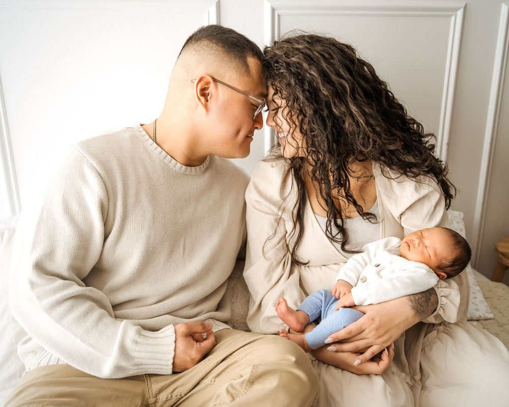 A couple shares a tender moment touching foreheads while cradling their newborn baby in soft neutral tones.