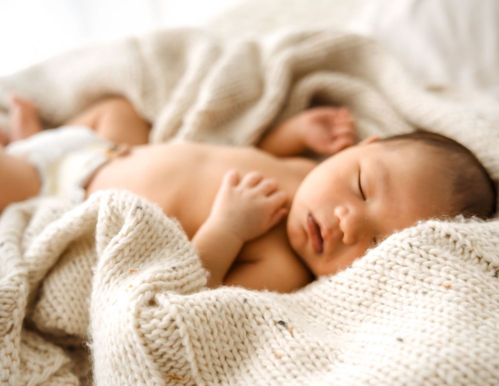 Two newborn babies sleeping peacefully on a soft knit blanket in a bright, serene setting.