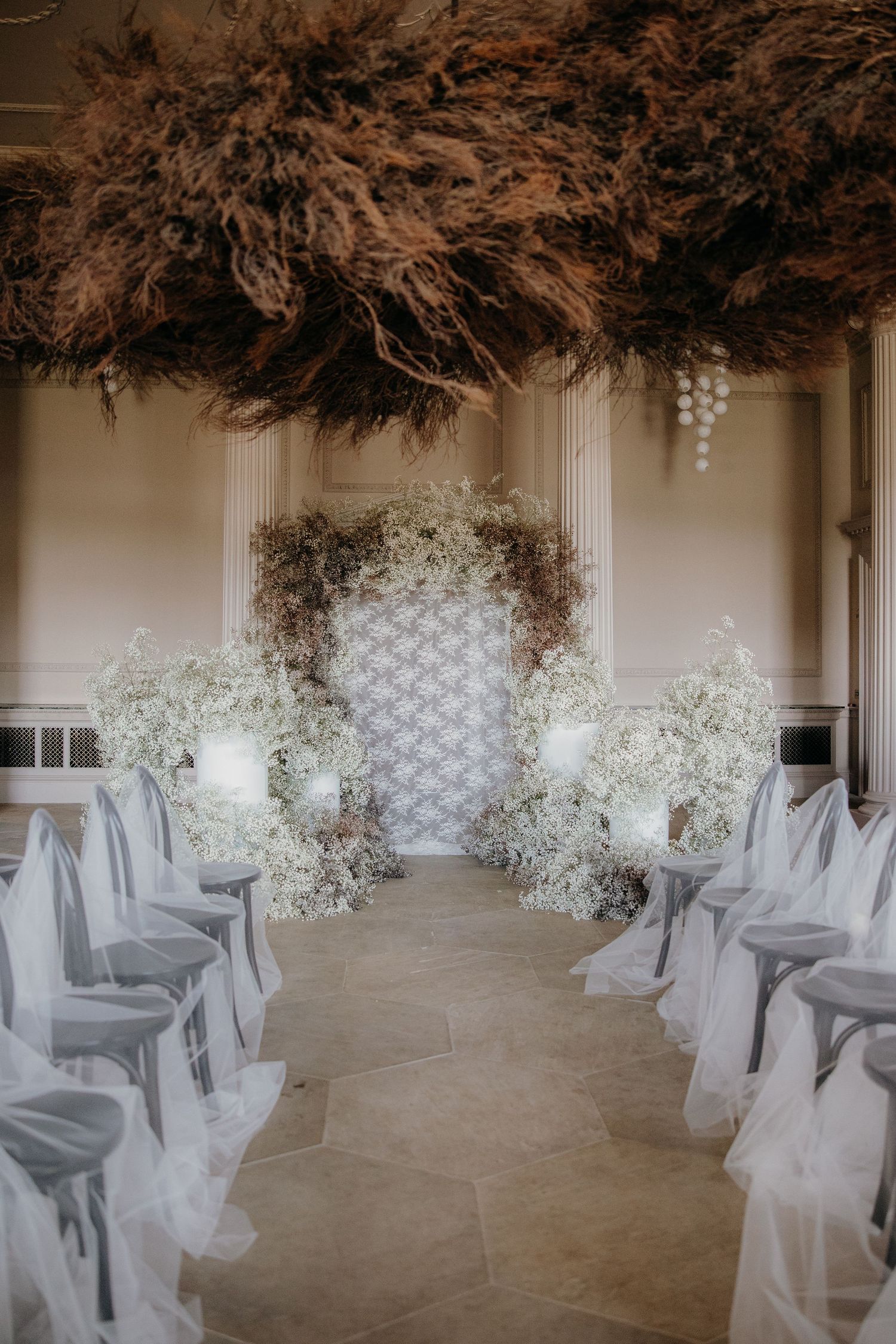 Elegant wedding ceremony aisle with white floral arrangements, draped chairs, and a decorative arch under dramatic dried pampas grass ceilin