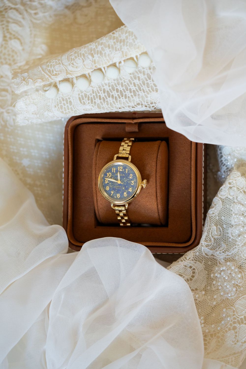 A gifted gold Shinola watch from parents to bride. The tan Shinola watch box is surrouned by an ivory vintage inspired wedding dress