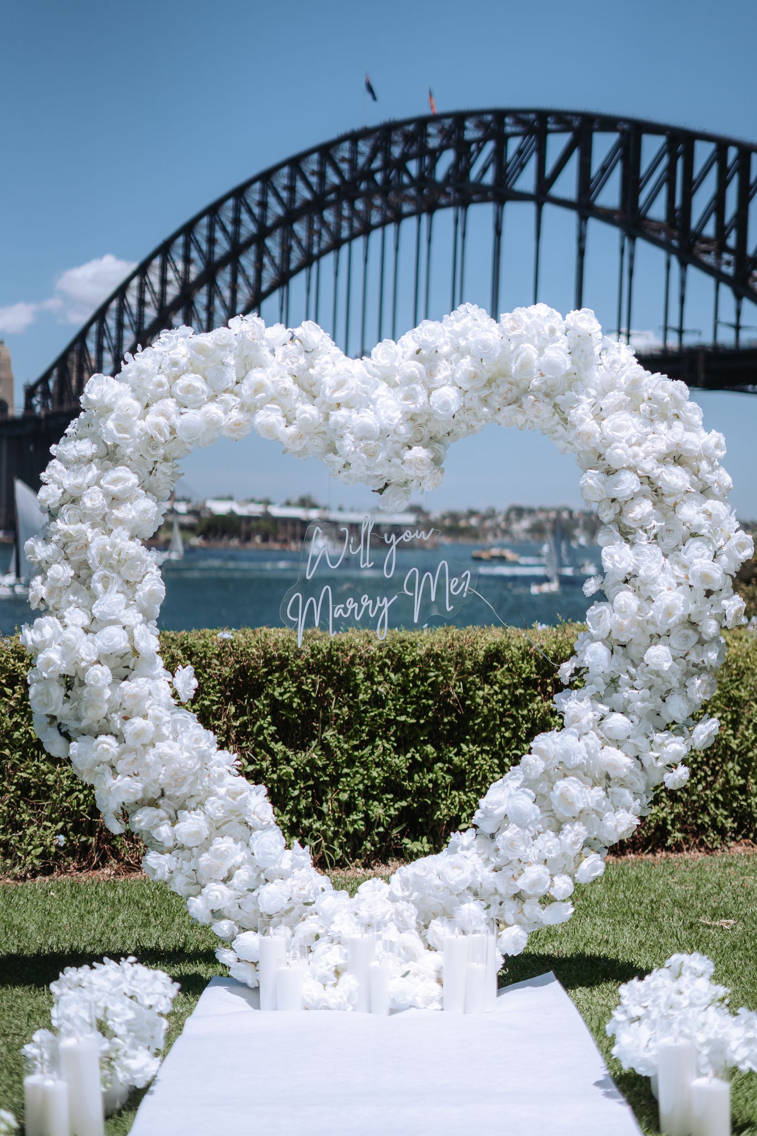 Heart-shaped white floral arch with Will you Marry Me sign, Sydney Harbour Bridge backdrop, outdoor wedding proposal setup.