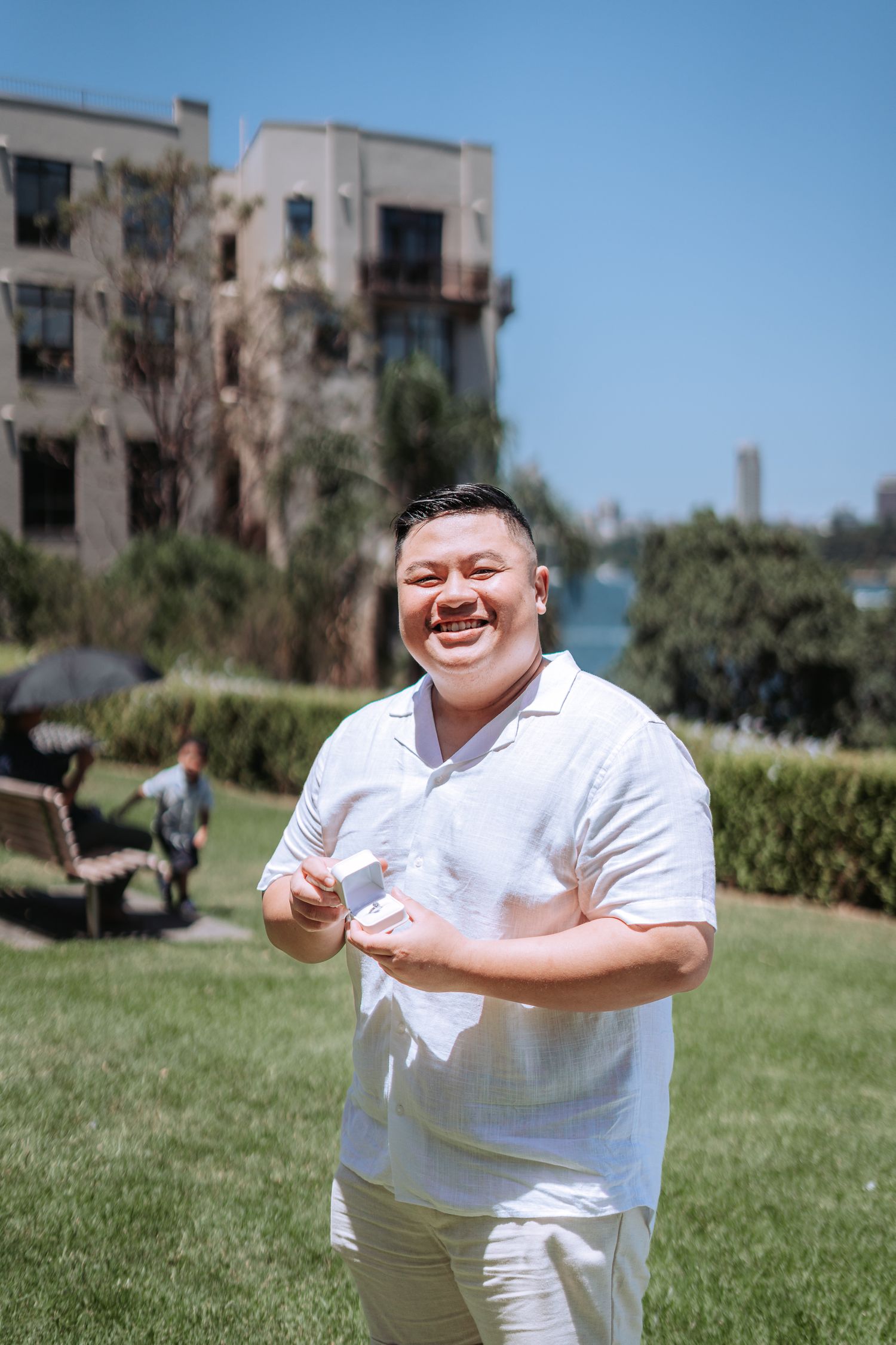 Smiling Asian man in white shirt holding a ring box on a sunny day in a park with a building in the background.