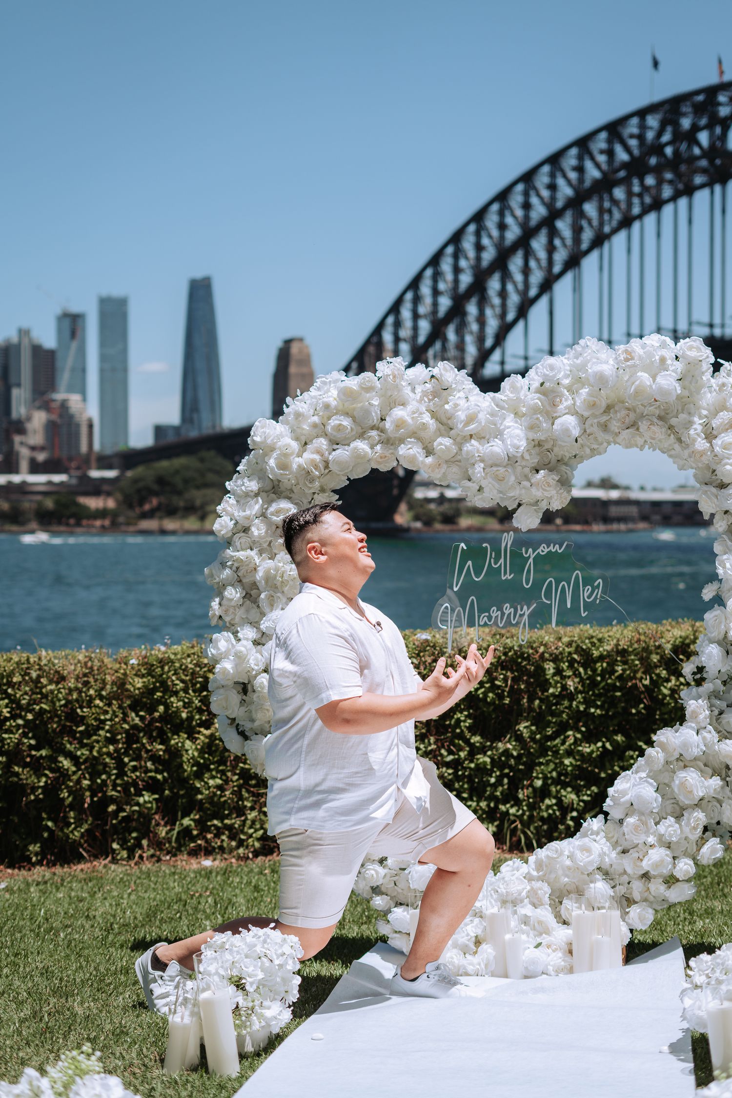 Man in white kneeling on one knee under a floral heart arch with Will You Marry Me sign, Sydney Harbour Bridge backdrop.