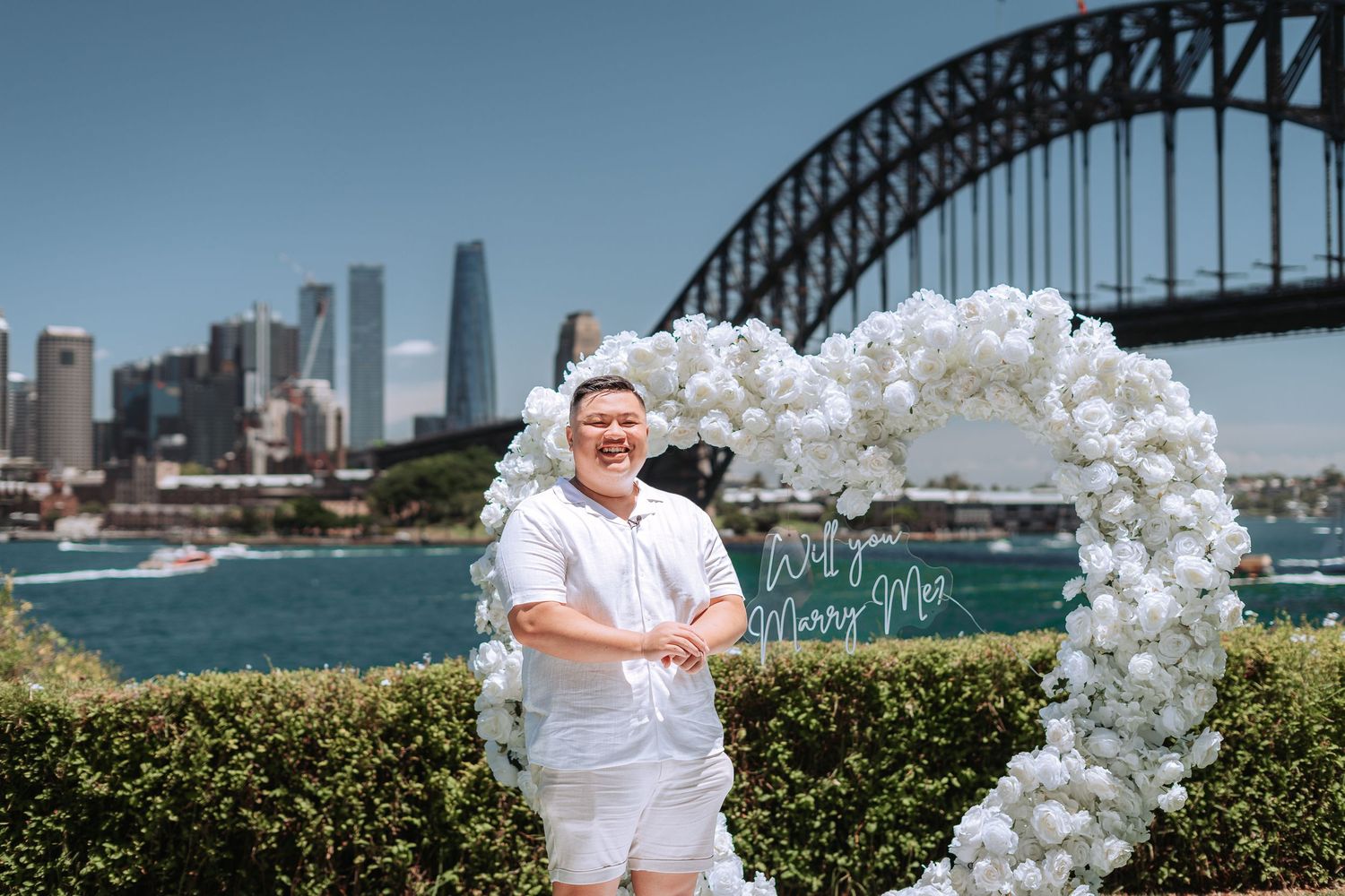 Man in white outfit smiling beside a white floral heart arch with Will You Marry Me sign near Sydney Harbour Bridge.