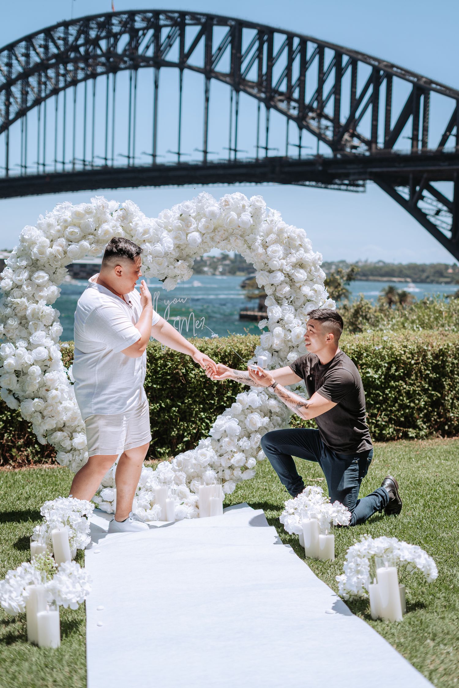 Man proposes on one knee under white floral heart arch with Sydney Harbour Bridge backdrop.