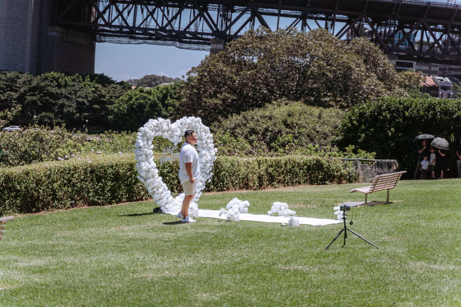 Person arranging a white balloon heart arch on green lawn near Sydney Harbour Bridge for an outdoor wedding ceremony.