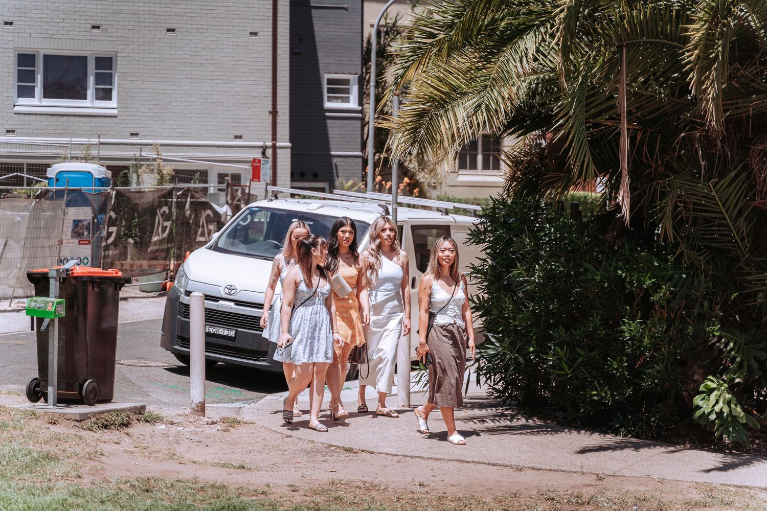 Group of women in summer dresses walking together on a sunny suburban street near a white van and palm trees.