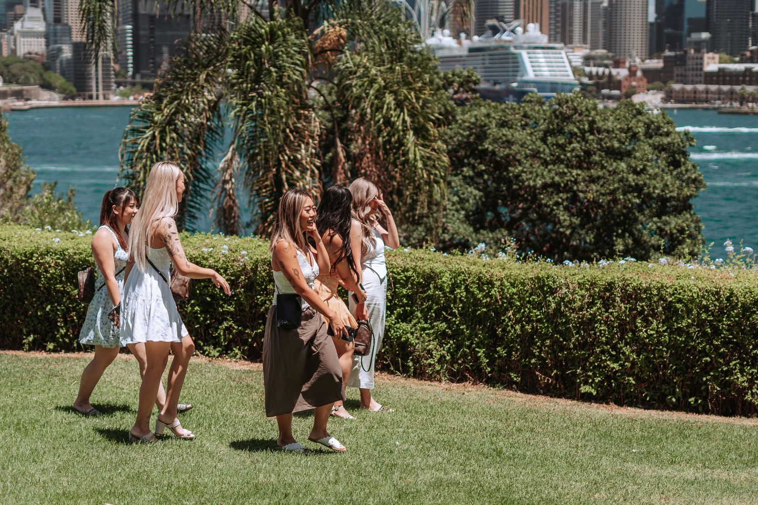 Group of women in summer dresses walking on grass near Sydney Harbour waterfront on a sunny day.