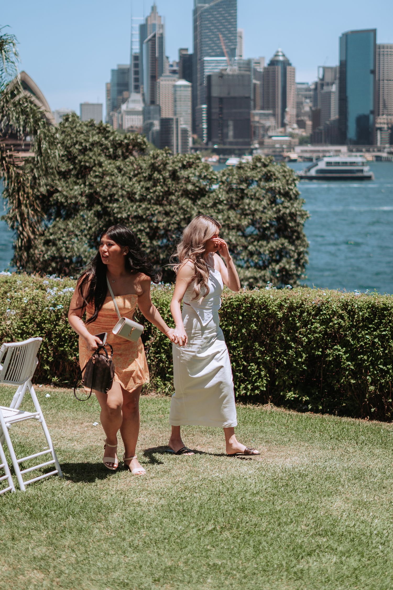 Two women in summer dresses hold hands on a grassy lawn with Sydney CBD skyline and harbour in background.