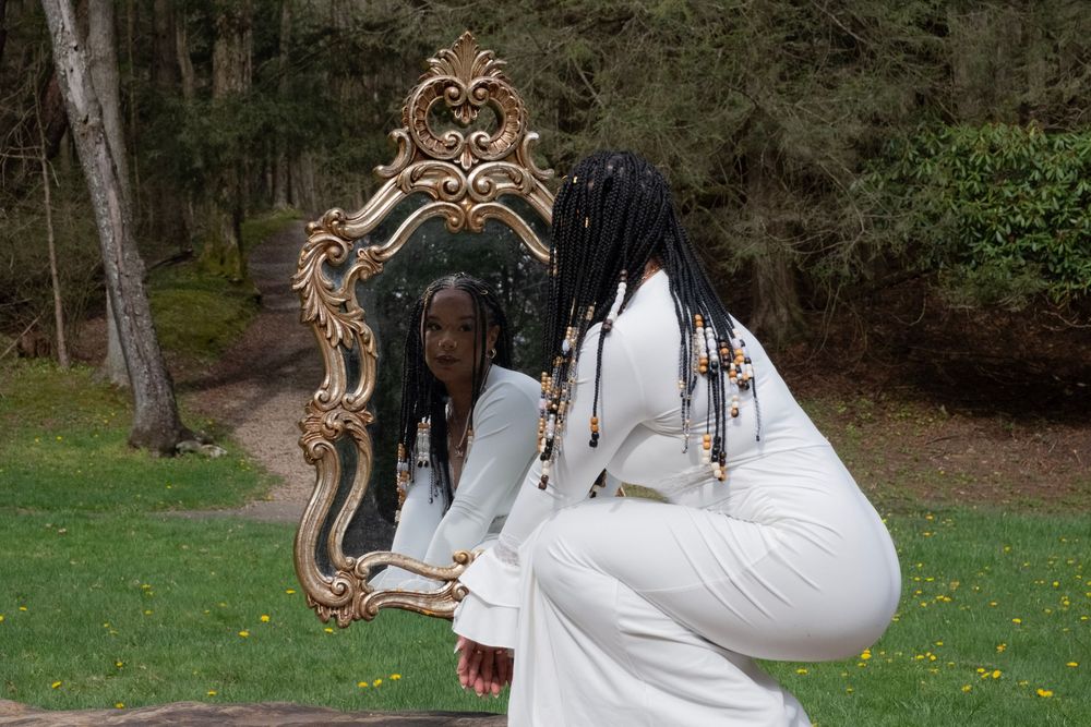 Woman in white outfit gazes into ornate gold mirror outdoors, surrounded by lush green trees.

Uniontown, PA