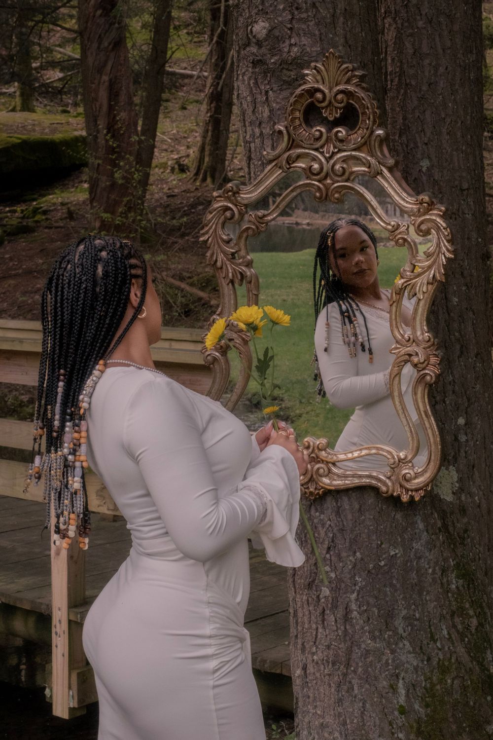 A Black woman in a white dress with braids holds yellow flowers while gazing into an ornate gold mirror outdoors.
Uniontown, PA