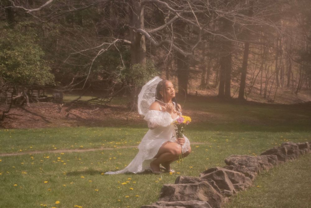 Bride in white wedding dress and veil kneeling on stone wall holding flowers in a lush green forest setting.
Uniontown, PA
