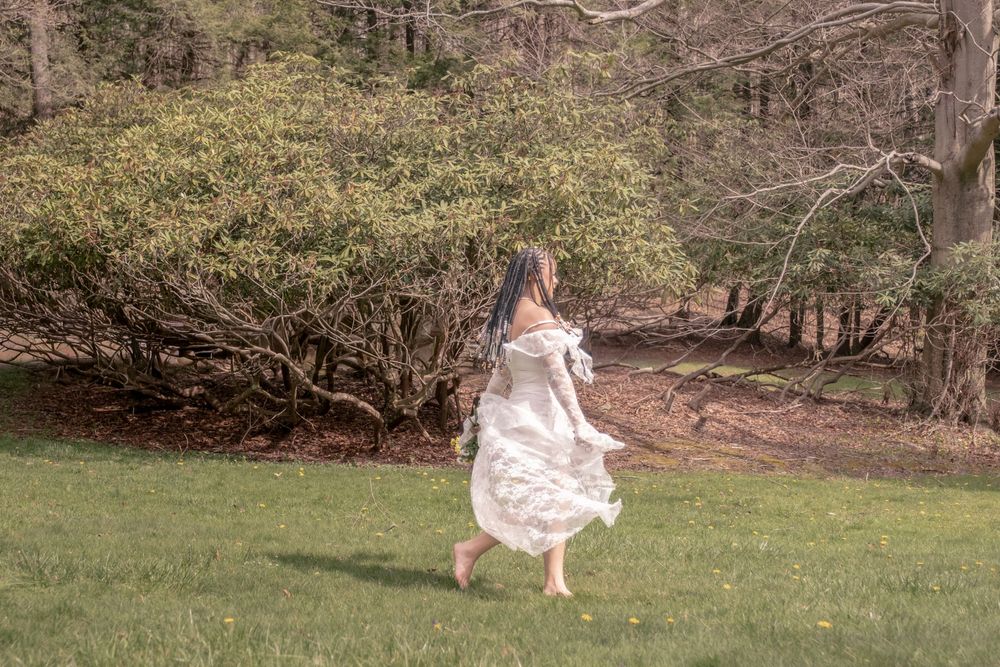 A barefoot woman in a flowing white dress runs across a green lawn near lush shrubs and trees.

Uniontown, PA
