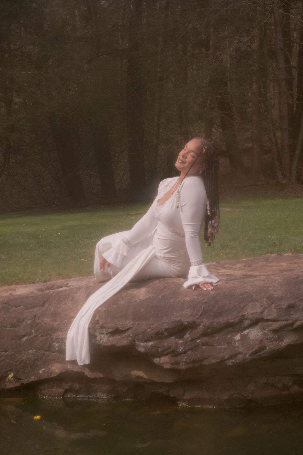 Woman in elegant white maxi dress sits serenely on a rock by a pond surrounded by lush green forest.

Uniontown, PA
