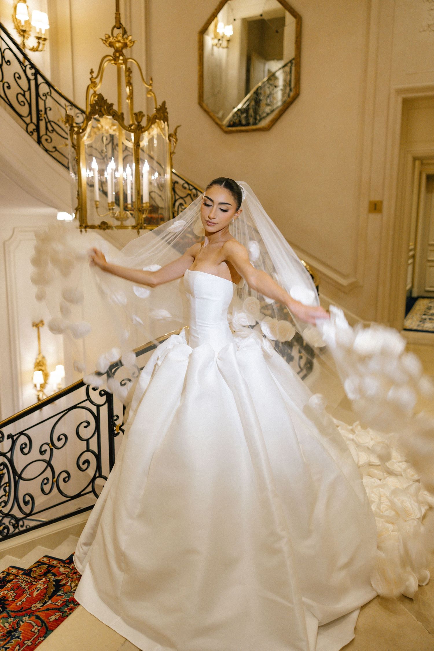 A couple walks down an outdoor aisle lined with flowers and cypress trees leading to an elegant villa entrance.