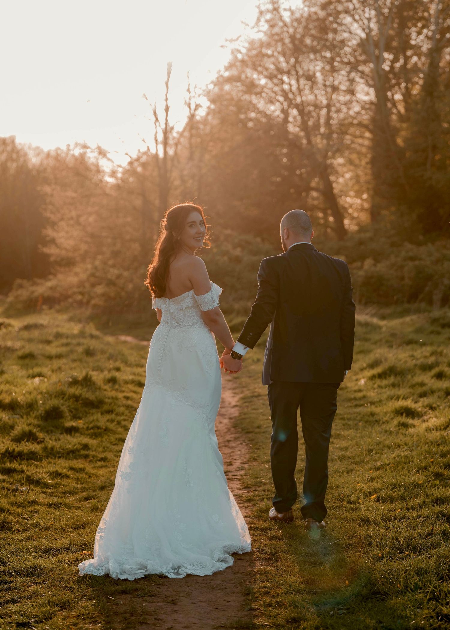 Bride in white off-shoulder gown holds groom's hand walking toward golden sunset in lush green field.
