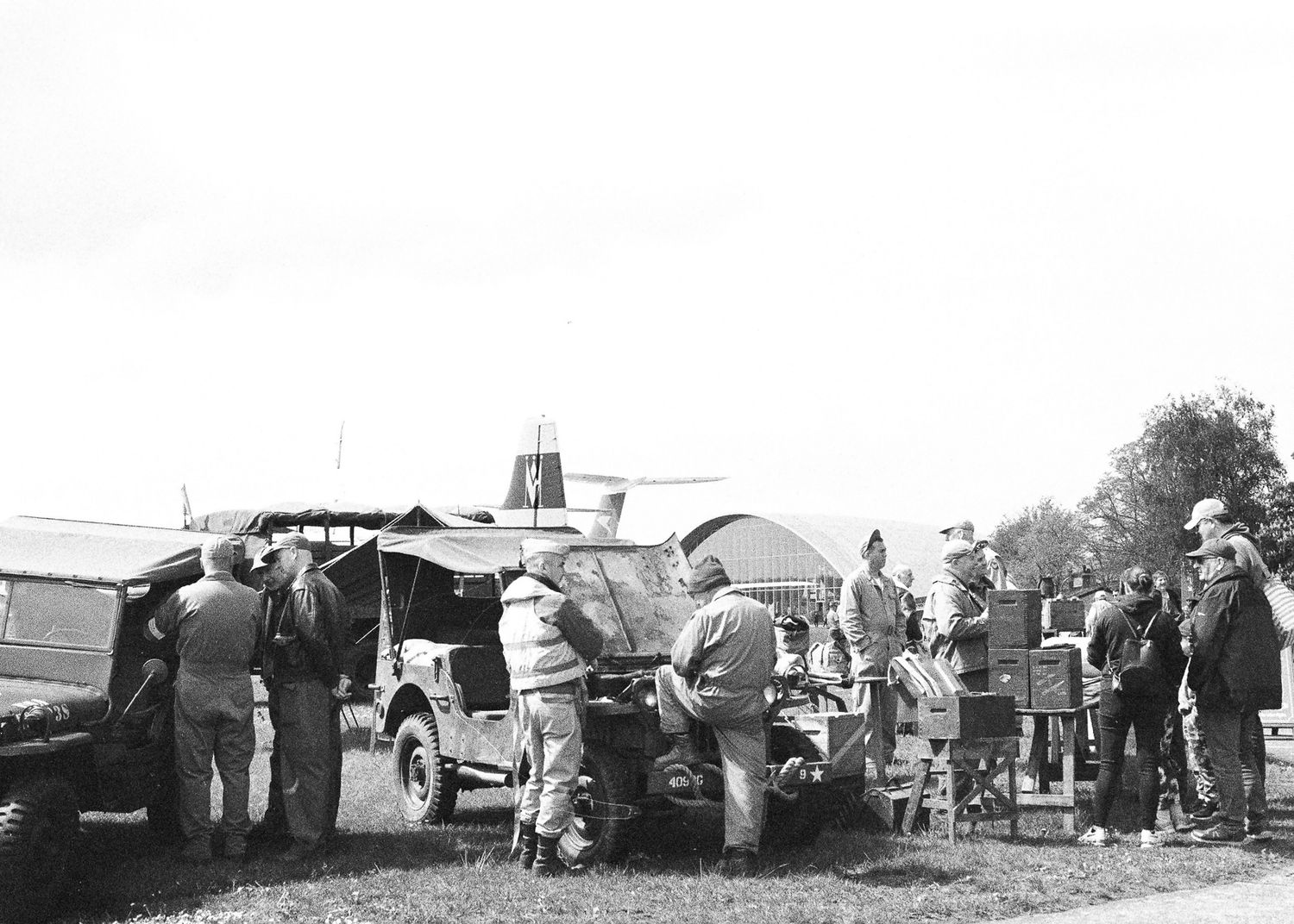 Black and white photo of people gathered around military vehicles in an open field during a historical reenactment event.