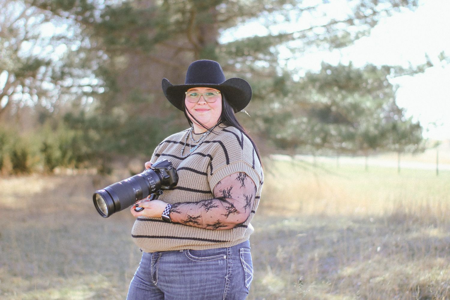 Woman in black cowboy hat holding a professional camera lens outdoors in a field with trees in background.