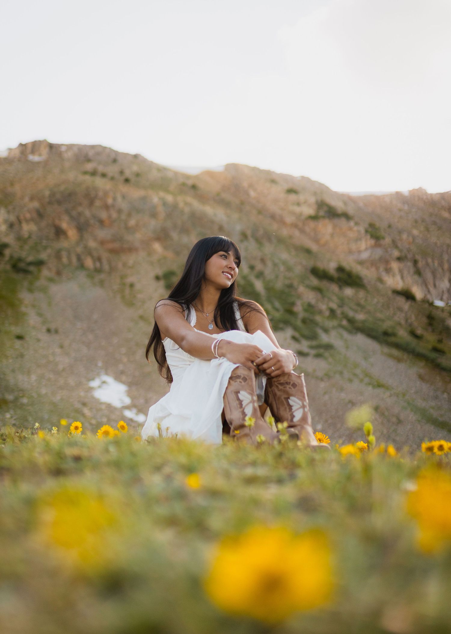 A smiling woman in a white dress and cowboy boots sits in a wildflower meadow with golden hills behind her.