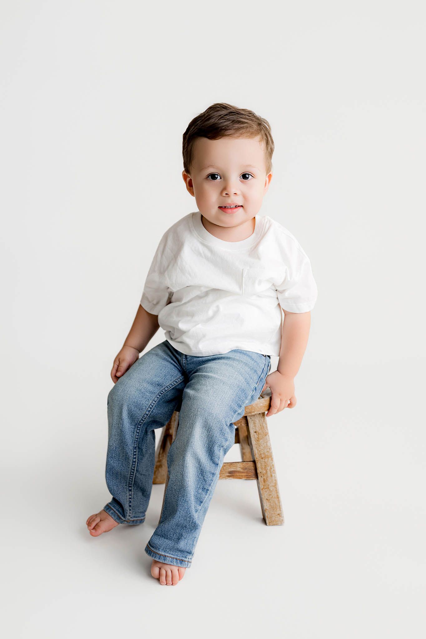 A cute toddler boy sits on a small wooden stool wearing a white t-shirt and jeans against a white background for 2nd birthday photos.